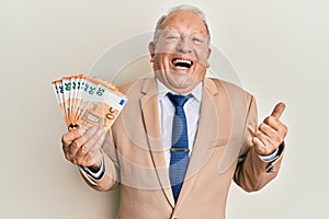Senior caucasian man holding euro banknotes pointing thumb up to the side smiling happy with open mouth