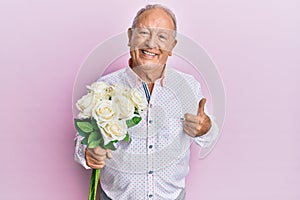 Senior caucasian man holding flowers smiling happy and positive, thumb up doing excellent and approval sign