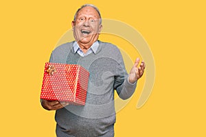 Senior caucasian man holding gift celebrating victory with happy smile and winner expression with raised hands