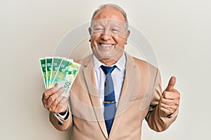 Senior caucasian man holding israel shekels smiling happy and positive, thumb up doing excellent and approval sign