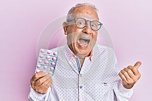 Senior caucasian man holding pills pointing thumb up to the side smiling happy with open mouth