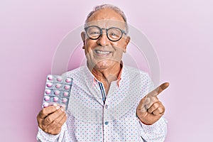 Senior caucasian man holding pills smiling happy pointing with hand and finger to the side