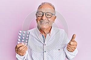 Senior caucasian man holding pills smiling happy and positive, thumb up doing excellent and approval sign