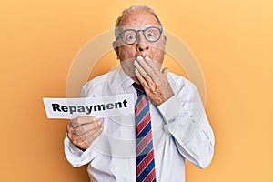 Senior caucasian man holding repayment word paper covering mouth with hand, shocked and afraid for mistake