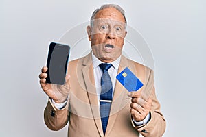 Senior caucasian man holding smartphone and credit card in shock face, looking skeptical and sarcastic, surprised with open mouth