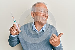 Senior caucasian man holding syringe pointing thumb up to the side smiling happy with open mouth