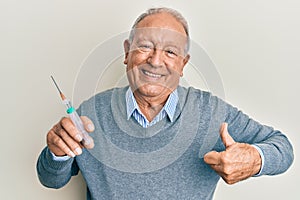 Senior caucasian man holding syringe smiling happy and positive, thumb up doing excellent and approval sign