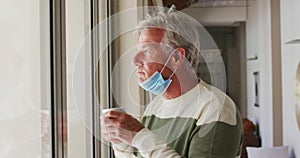 Senior caucasian man with lowered face mask drinking coffee while looking out of window at home