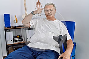 Senior caucasian man at physiotherapy clinic holding crutches smiling pointing to head with one finger, great idea or thought,