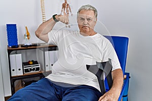 Senior caucasian man at physiotherapy clinic holding crutches strong person showing arm muscle, confident and proud of power