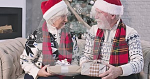 Senior Caucasian man and positive woman sitting on sofa and holding Christmas presents. Husband looking at his adorable