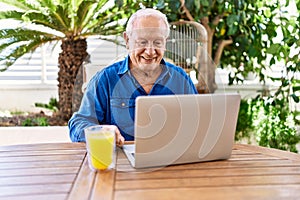 Senior caucasian man using laptop and drinking orange juice sitting on the table at terrace