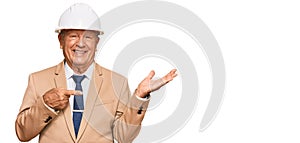 Senior caucasian man wearing architect hardhat amazed and smiling to the camera while presenting with hand and pointing with