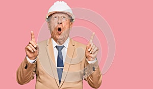 Senior caucasian man wearing architect hardhat amazed and surprised looking up and pointing with fingers and raised arms