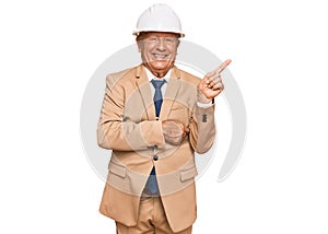 Senior caucasian man wearing architect hardhat smiling happy pointing with hand and finger to the side