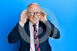 Senior caucasian man wearing business suit and tie trying to hear both hands on ear gesture, curious for gossip