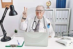 Senior caucasian man wearing doctor uniform and stethoscope at the clinic smiling looking to the camera showing fingers doing