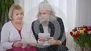 Senior Caucasian man and woman in pajamas watching old photos sitting on bed at home. Relaxed couple recalling memories