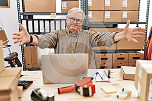 Senior caucasian man working at small business ecommerce with laptop looking at the camera smiling with open arms for hug