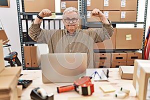 Senior caucasian man working at small business ecommerce with laptop showing arms muscles smiling proud