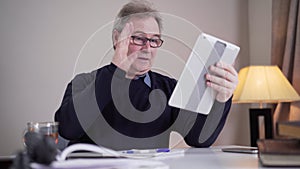 Senior Caucasian man writing at the table when having incoming video call. Mature male retiree in eyeglasses waving and