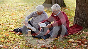 Senior Caucasian married couple read together on the ground in the park full shot selective focus