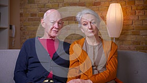 Senior caucasian spouses watch news on TV being attentive at sofa in living room.