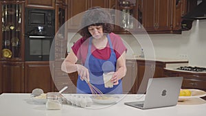 Senior Caucasian woman checking recipe online and mixing milk into dough. Housewifely retiree cooking new bakery at home