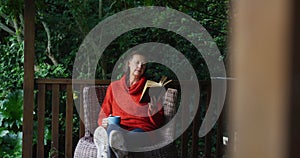 Senior caucasian woman relaxing in garden, sitting with feet up, drinking coffee and reading book