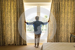 Senior caucasian woman standing next to window and widening courtains in bedroom