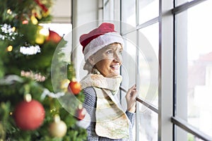 Senior Caucasian woman wearing red Santa hat looking out the window in white snow during christmas holiday