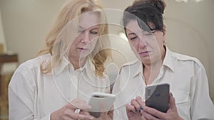 Senior Caucasian women teaching her friend using smart phone. Portrait of two mature ladies in white shirts spending