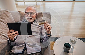 Senior cheerful handsome man lying down on sofa or couch at his home or office using hand held tablet device and credit card for