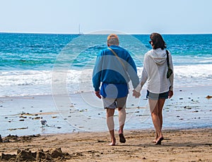 Senior citizen baby boomer male and female caucasian couple walking on the beach towards the ocean holding hands.