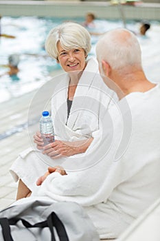 senior couple in bathrobes sat resting by pool