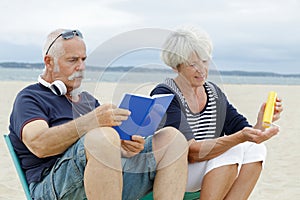 senior couple on beach reading book and applying suncream