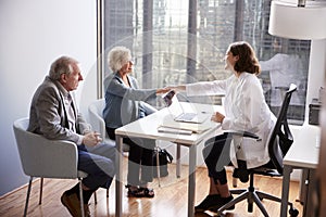 Senior Couple Being Greeted By Female Doctor With Handshake On Visit To Hospital For Consultation