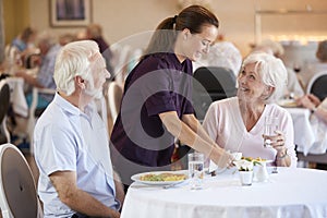 Senior Couple Being Served With Meal By Carer In Dining Room Of Retirement Home