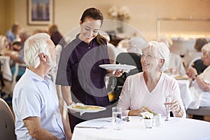 Senior Couple Being Served With Meal By Carer In Dining Room Of Retirement Home