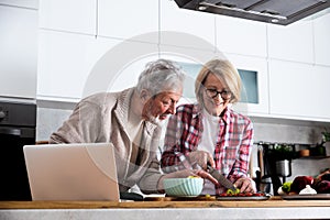 Senior couple cooking dinner together in the kitchen for golden wedding anniversary, reading recipe from internet on laptop. Older