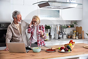 Senior couple cooking dinner together in the kitchen for golden wedding anniversary, reading recipe from internet on laptop. Older