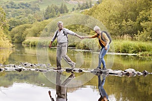 Senior Couple Crossing River Whilst Hiking In UK Lake District