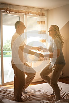 Senior couple dancing and jumping together on bed holding hands