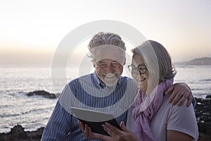 A senior couple embrace near the beach while looking at the tablet together. Sitting on the cliffs, twilight, two elderly people