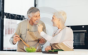 Senior Couple Feeding Each Other And Laughing Enjoying Cooking Indoors