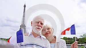 Senior couple with French flags near Eiffel Tower doing selfie. Smiling tourist traveling in Europe.
