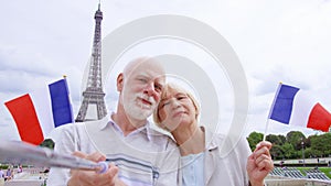Senior couple with French flags near Eiffel Tower doing selfie. Smiling tourist traveling in Europe.
