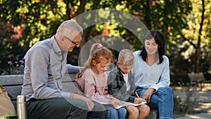Senior couple with grandchildren sitting on bench and doing homewrok outdoors in park.