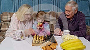 Senior couple grandfather, grandmother resting on sofa, playing chess with granddaughter child kid