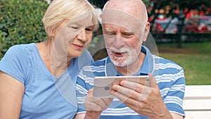 Senior couple having great time sitting on bench in park chatting relaxing, browsing in smartphone.